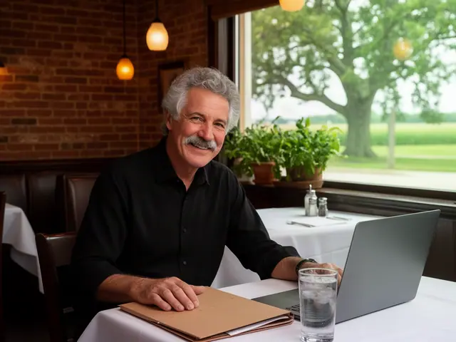 Smiling man with grey curly hair and a moustache at a restaurant table with a laptop and kraft folder